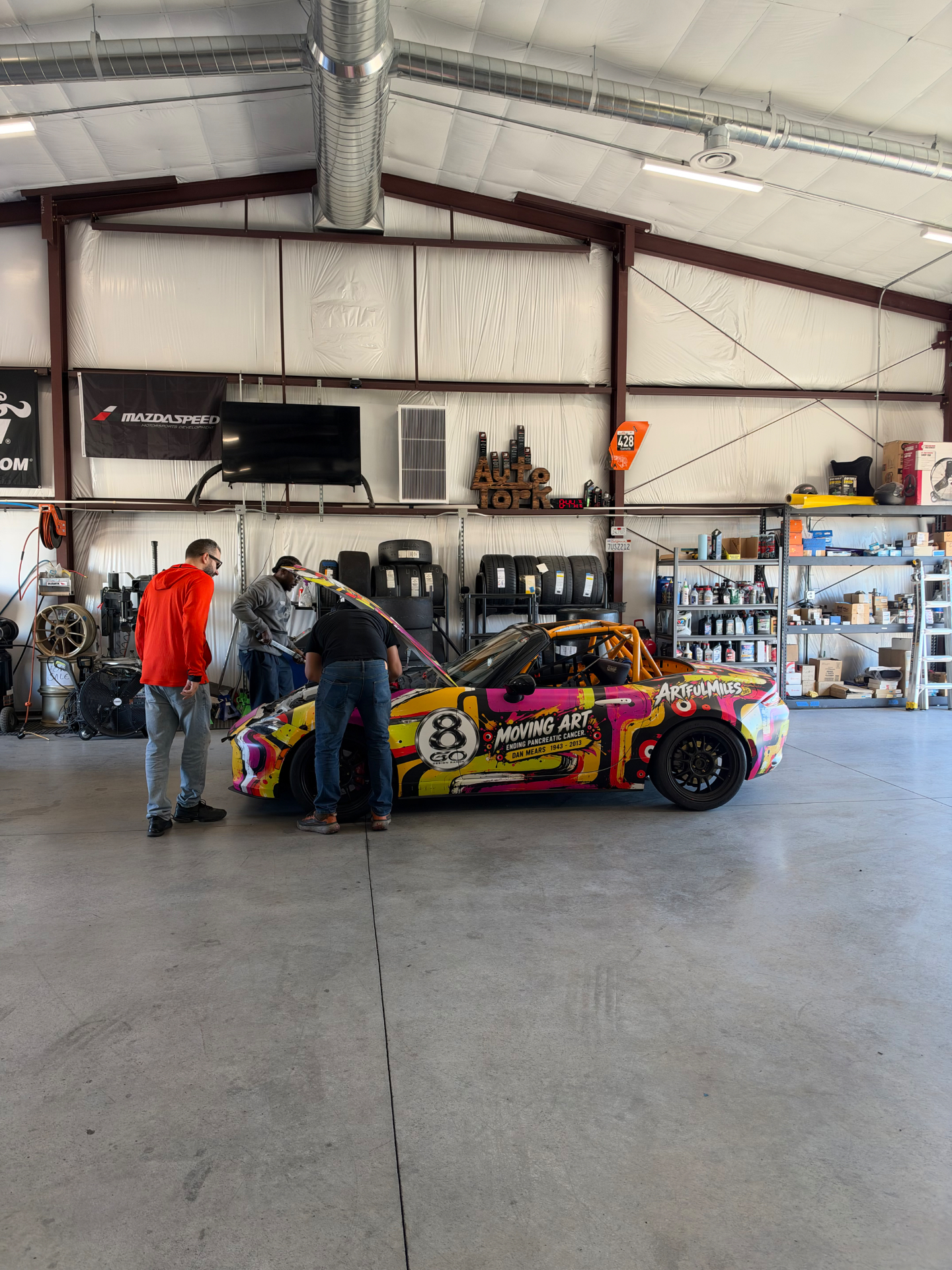 People gathered around an ArtfulMiles wrapped race car with its hood open in a workshop