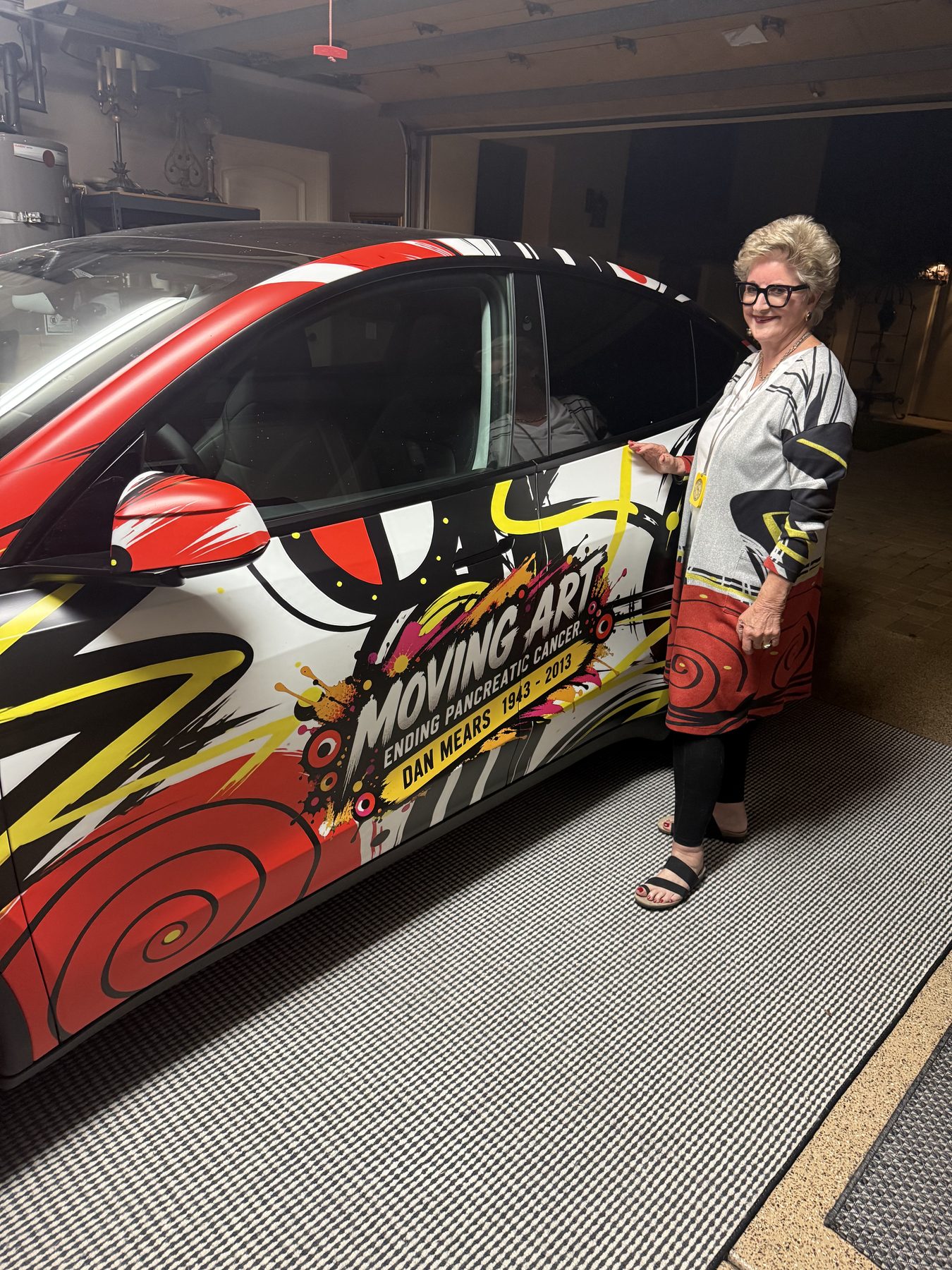 Susan Fielder standing beside a Tesla wrapped in red, yellow and black Moving Art artwork inside a garage