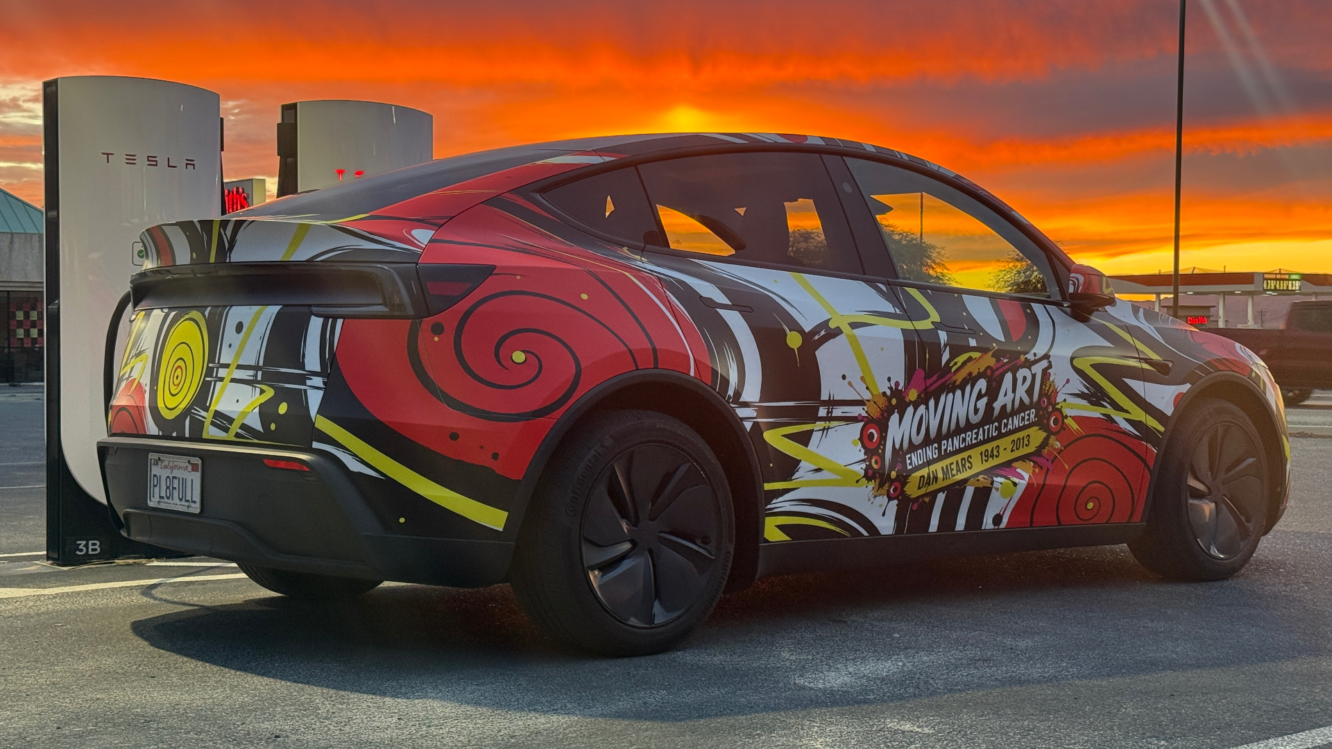 Tesla at a charging station under an orange sunset, wrapped in red spirals, yellow accents and a Moving Art tribute to Dan Mears